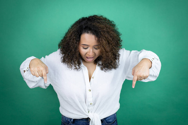Jeune fille afro-américaine portant une chemise blanche sur fond vert surpris, regardant vers le bas et pointant vers le bas avec les doigts et les bras levés - Photo, image