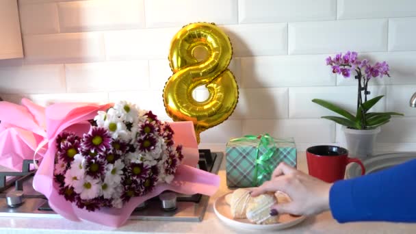 Жінка бере печиво на кухні з золотою кулькою 8, весняними квітами, подарунковою коробкою і сніданком. День матері. 8 березня - Міжнародний жіночий день - Кадри, відео