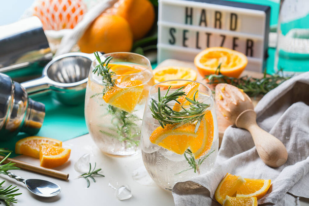 Harter Seltzer-Cocktail mit Orange, Rosmarin und Eis auf einem Tisch. Sommer-Erfrischungsgetränk, Drink mit trendigen Zero-Waste-Accessoires, Bambusstroh und Netztasche. - Foto, Bild
