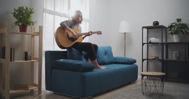 The girl plays the guitar on the couch. A woman alone creates music in a white living room. The artist plays the classical guitar. The musician composes a dreary melody. Long short. - Metraje, vídeo