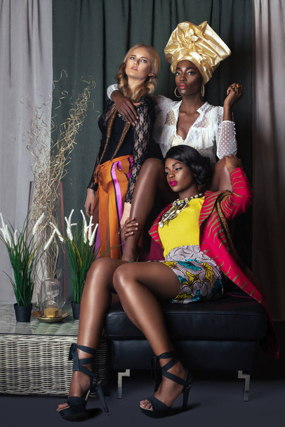 Portrait of a group of 3 elegant women with curly long hair and beautiful makeup posing by themselves in a studio with curtain background wearing designer clothing and high heels. - Photo, Image