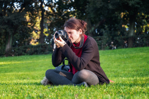Menina sentada na grama do parque público, fotografando a paisagem circundante com sua câmera reflexa. Casual, morena. - Foto, Imagem