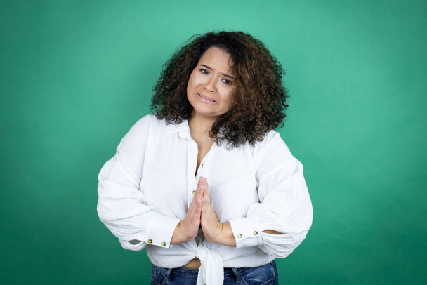 Νεαρή Αφροαμερικανή που φοράει λευκό πουκάμισο πάνω από πράσινο φόντο ικετεύει και προσεύχεται με τα χέρια μαζί με την έκφραση της ελπίδας στο πρόσωπο πολύ συναισθηματική και ανησυχούν - Φωτογραφία, εικόνα