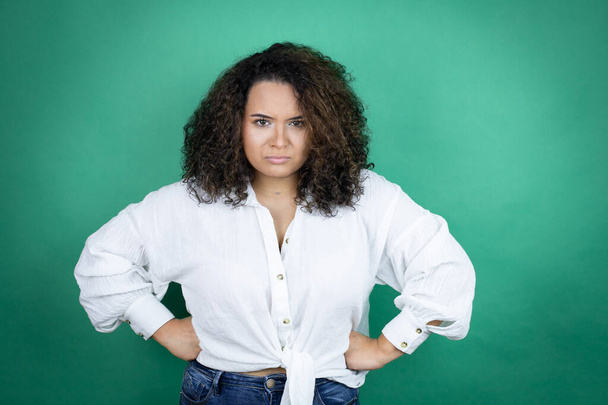 Jeune fille afro-américaine portant une chemise blanche sur fond vert sceptique et nerveux, expression désapprobatrice sur le visage avec les bras à la taille - Photo, image