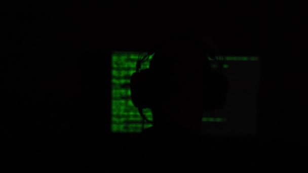 Hacker seduto di fronte a uno schermo del computer con codice verde in una stanza buia e ombreggiata. Profondità di campo ridotta. - Filmati, video