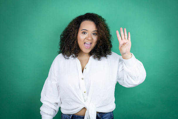 Jeune fille afro-américaine portant chemise blanche sur fond vert montrant et pointant vers le haut avec les doigts numéro quatre tout en souriant confiant et heureux - Photo, image