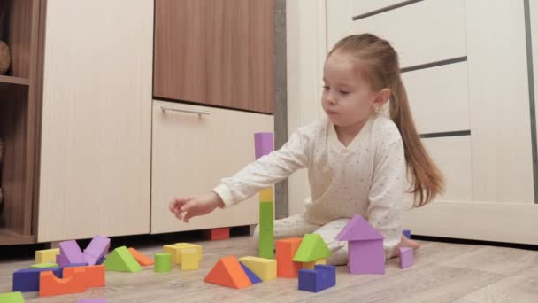 Lapsi tyttö leikkii värillisiä kuutioita lastenhuoneessa lattialla. Poika rakentaa omakotitaloa. Opetuspelit lasten kehittämiseen. Lapsen opettaminen leikin kautta. Onnellinen perhe, tytär - Materiaali, video