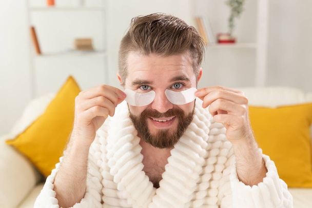 Een man met een baard verwijdert oogvlekken op zijn gezicht. Rimpels en gezicht thuiszorg voor mannen. - Foto, afbeelding