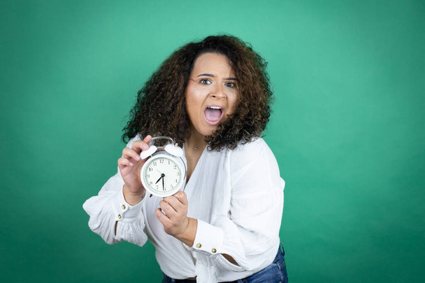Giovane ragazza afroamericana indossa camicia bianca su sfondo verde urla e paura, indicando l'orologio - Foto, immagini