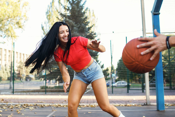 Una bella giovane bruna sta giocando a basket con il suo ragazzo e sta cercando di prendere la palla. La coppia sportiva si sta divertendo e godendo il processo. Ragazza in pantaloncini di jeans e una t-shirt rossa - Foto, immagini