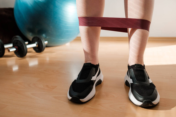 Spor tozluklu kadın bacakları ve spor ayakkabıları kilitlenme sırasında evde spor lastiğiyle egzersiz yapıyor. Ev antrenmanı. direnç bantları ile.  - Fotoğraf, Görsel