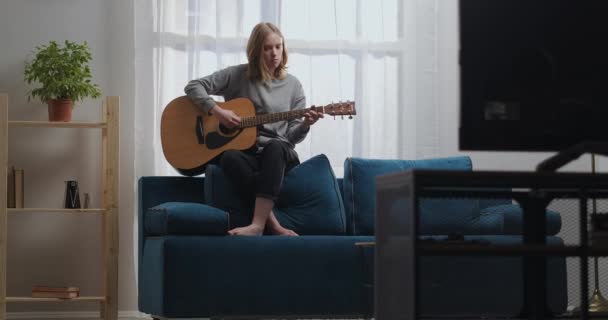 Teini-ikäinen tyttö harmaassa puserossa ja mustat housut ilman sukkia soittaa rauhallista melodiaa akustisella kitaralla. Istuu sinisen sohvan takaosassa. Aurinko paistaa ikkunasta takana. - Materiaali, video