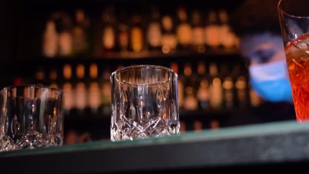 Barman versando whisky dorato, cognac o brandy dalla bottiglia in vetro con cubetti di ghiaccio sul tavolo di legno. Oro lucido partito celebrazione sfondo. - Filmati, video