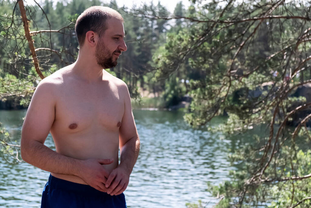 Бородатий без сорочки чоловік проводить час на озері. Він стоїть на тлі води і дерев в спекотний літній день
 - Фото, зображення