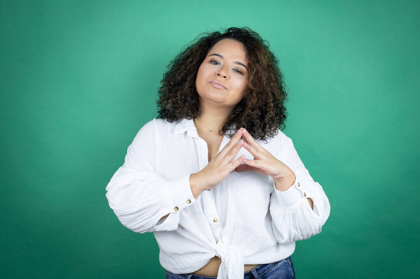 Young african american girl wearing white shirt over green background with Hands together and fingers crossed smiling relaxed and cheerful. Success and optimistic - Photo, Image