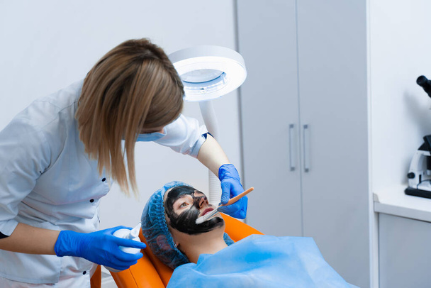 Dermatologist smears black mask on face for laser photorejuvenation and carbon peeling. Dermatology and cosmetology. Using surgical laser. - Photo, Image