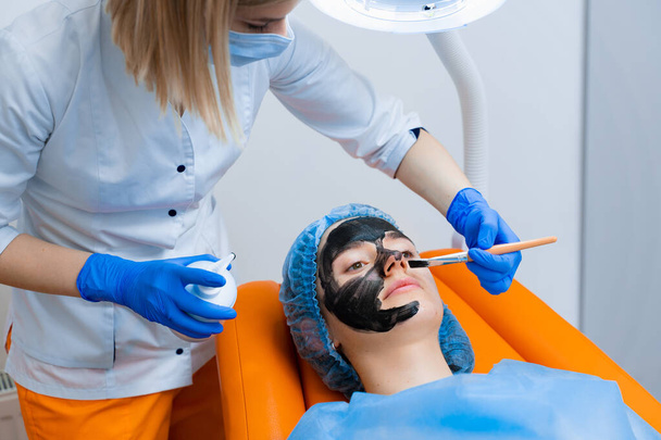 Dermatolog rozmazuje czarną maskę na twarzy do laserowej fotorejuvenacji i obierania węgla. Dermatologia i kosmetologia. Używanie lasera chirurgicznego. - Zdjęcie, obraz