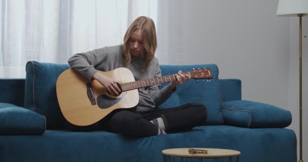 Ένα έφηβο κορίτσι στο σπίτι ρούχα παίζει μια νέα μελωδία μελωδία σε μια κλασική κιθάρα κάθεται σε μια άνετη θέση σε ένα μπλε καναπέ σε ένα ζεστό σαλόνι - Πλάνα, βίντεο