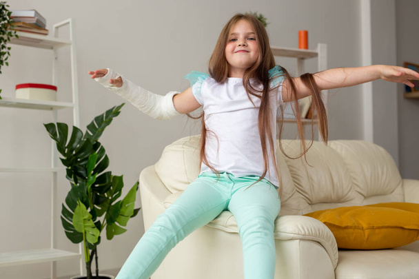 Gyermek gipsszel a törött csuklóján vagy karján mosolyog és szórakozik a kanapén. Pozitív hozzáállás, felépülés és gyerekkoncepció. - Fotó, kép