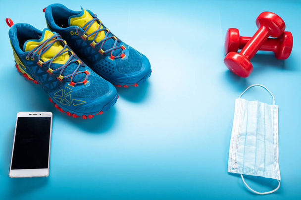 Αθλητικά αθλητικά παπούτσια, smartphone και ιατρική μάσκα σε μπλε φόντο. Υποχρεωτικά πράγματα στο γυμναστήριο. Όψη γωνίας. - Φωτογραφία, εικόνα