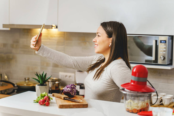 Mutfakta selfie çekerken gülümseyen yetişkin beyaz kadın portresi ya da evde yemek hazırlarken telefon görüşmesi yapan gerçek insanlar konsepti. - Fotoğraf, Görsel