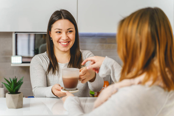 Happy caucasian woman smile while giving cup of coffee or hot chocolate to her unknown female friend while standing at the kitchen at home - real people friendship concept - Photo, Image