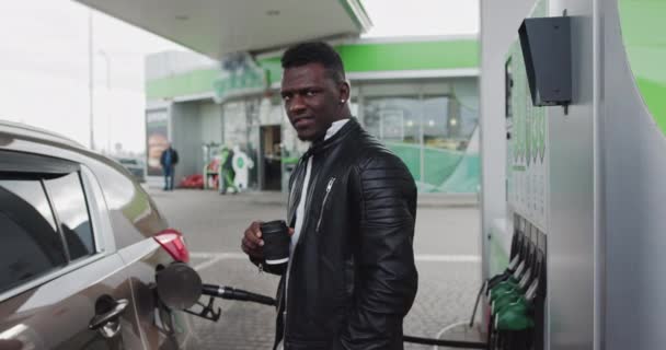 Porträt eines Afrikaners an einer Tankstelle mit Kaffee in der Hand. Während der Fahrt das Auto betanken, am GAS-Bahnhof anhalten, Kaffee trinken. Kraftstoff, Tankstelle, Benzinpreiskonzept - Filmmaterial, Video