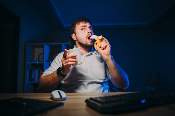 Konsantre olmuş serbest programcı geceleri evde bilgisayar başında oturup muz ve çayla yemek yiyor. Sakallı adam bilgisayarla uğraşırken geceleri meyve yiyor.. - Fotoğraf, Görsel