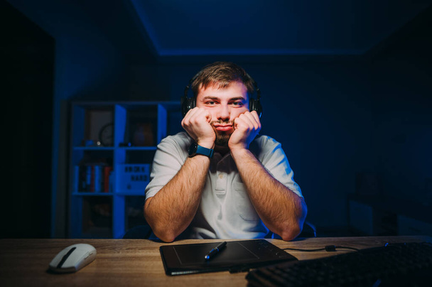 Een overstuur bebaarde man werkt 's nachts thuis op de computer, kijkt naar het scherm en valt in slaap. Trieste freelancer ontevreden met het werken 's nachts op de computer. - Foto, afbeelding
