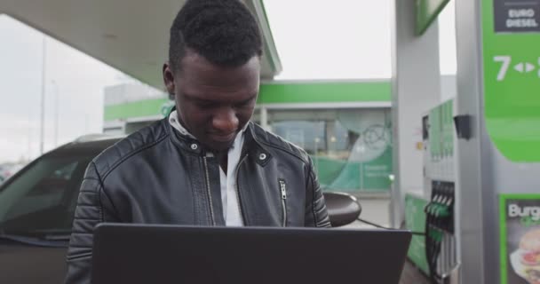 Ein afroamerikanischer Freiberufler hält einen Laptop in der Hand und lehnt sich an ein Luxusauto. Arbeit unterwegs, Freelancer. Auto an der Tankstelle. Kraftstoff, Tankstelle, Benzinpreiskonzept. - Filmmaterial, Video
