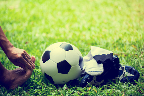 Bola de fútbol y zapatos deportivos en hierba verde con jugador de fútbol en el dolor de pies. Jugador de fútbol sentado porque el dolor de tobillo de jugar. El concepto de lesión deportiva. - Foto, imagen