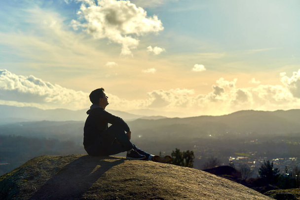 mladý chlapec sedící na vrcholu hory a hledící k obzoru. podnikavý muž sedí na skále s pozadí západu slunce krajiny. tmavý teenager s latinskoamerickými rysy na hoře - Fotografie, Obrázek
