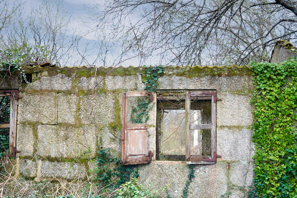 Opuszczony kamienny dom z rozbitym drewnianym oknem. stare okno z drewnem w starym galicyjskim domu z zachmurzonym niebem i roślinnością wokół niego. - Zdjęcie, obraz