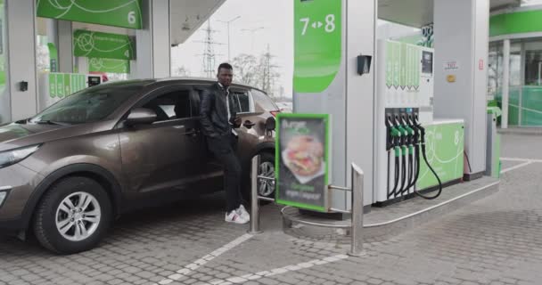 Porträt eines Afrikaners an einer Tankstelle mit Kaffee in der Hand. Während der Fahrt das Auto betanken, am GAS-Bahnhof anhalten, Kaffee trinken. Tankstellenübersetzung: ENERGIE IHRER BEWEGUNG - Filmmaterial, Video