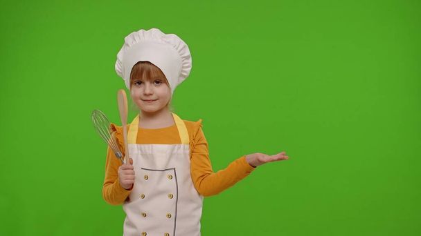 Enfant fille habillé cuisinier chef dans tablier pointant à droite sur l'espace vide sur fond chromatique clé - Photo, image
