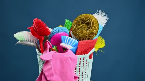 plastikowy kosz ze szczotkami, gąbkami i gumowymi rękawiczkami do czyszczenia pomieszczeń, obok butelki detergentu, niebieskie tło. Obiekty obracają się wokół własnej osi - Materiał filmowy, wideo