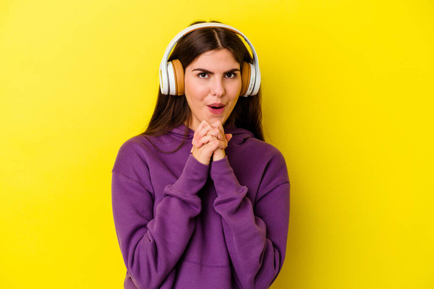 Jonge blanke vrouw luisteren naar muziek met koptelefoon geïsoleerd op roze achtergrond bidden voor geluk, verbaasd en opening mond op zoek naar voren. - Foto, afbeelding