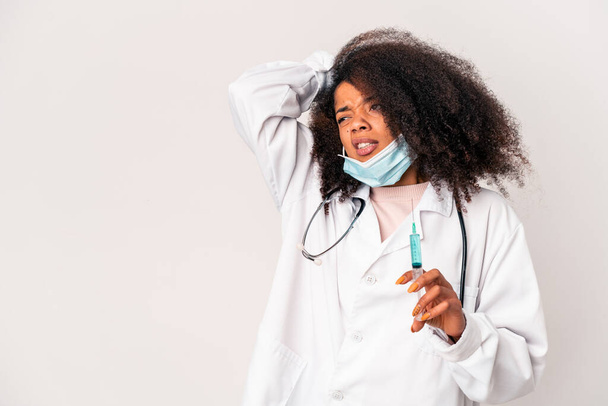 Νεαρή Αφροαμερικανή σγουρή γιατρός που κρατάει μια σύριγγα ακουμπώντας το πίσω μέρος του κεφαλιού, σκέφτεται και κάνει μια επιλογή. - Φωτογραφία, εικόνα