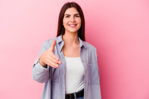 Young caucasian woman isolated on pink background stretching hand at camera in greeting gesture. - Fotoğraf, Görsel