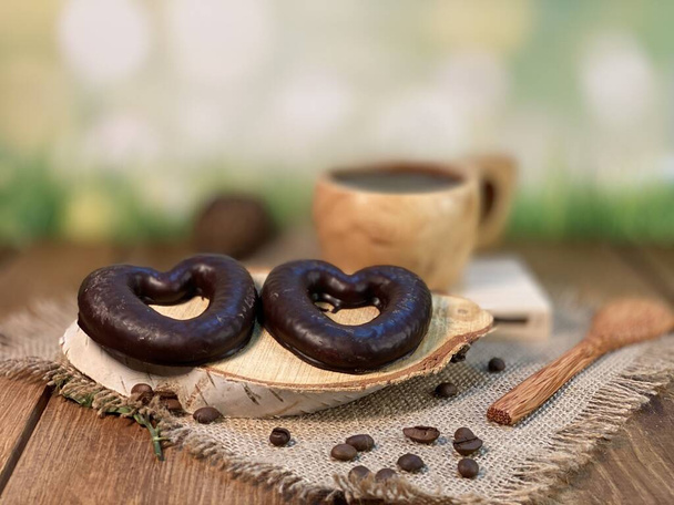 Oak table with visible grains, stand, pallet, on it there is a cup made of wood with black coffee. Coffee beans scattered on a jute napkin, a wooden spoon, background green grass and gingerbread heart is lying on a wooden pallet - Photo, Image