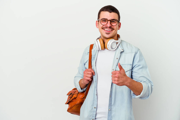 Joven estudiante caucásico escuchando música aislada sobre fondo blanco sonriendo y levantando el pulgar hacia arriba - Foto, imagen