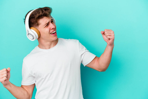 Jonge blanke man die naar muziek luistert met een koptelefoon geïsoleerd op blauwe achtergrond heffende vuist na een overwinning, winnaarsconcept. - Foto, afbeelding