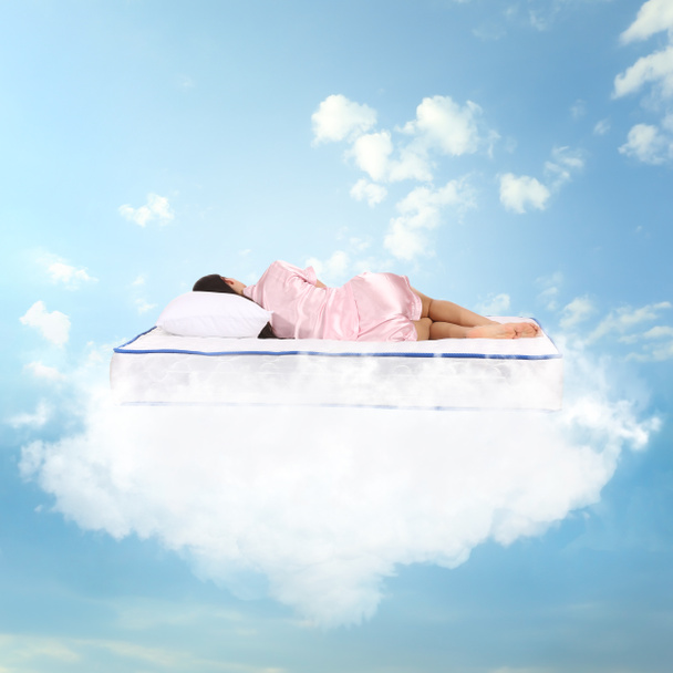 Νεαρή γυναίκα κοιμάται σε στρώμα απαλό σαν σύννεφα στον γαλάζιο ουρανό - Φωτογραφία, εικόνα