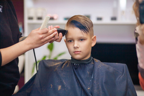 Um menino bonito senta-se em um cabeleireiro no estilista, uma criança em idade escolar está recebendo corte de cabelo em um salão de beleza, uma criança em uma barbearia, corte de cabelo de homens curtos. - Foto, Imagem