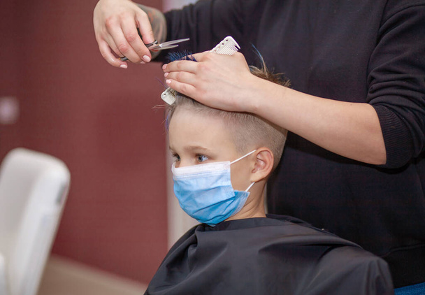 Un niño lindo con flequillo azul se sienta en una máscara protectora en una peluquería en el estilista, nuevo normal, cuarentena debido a la epidemia de covidio-19, un escolar se corta el pelo en un salón de belleza - Foto, Imagen
