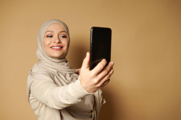 Piękny arabski kobieta z piękny ząb uśmiech i pokryte głowy podejmowania selfie stoi na beżowym tle z miejsca kopiowania. - Zdjęcie, obraz