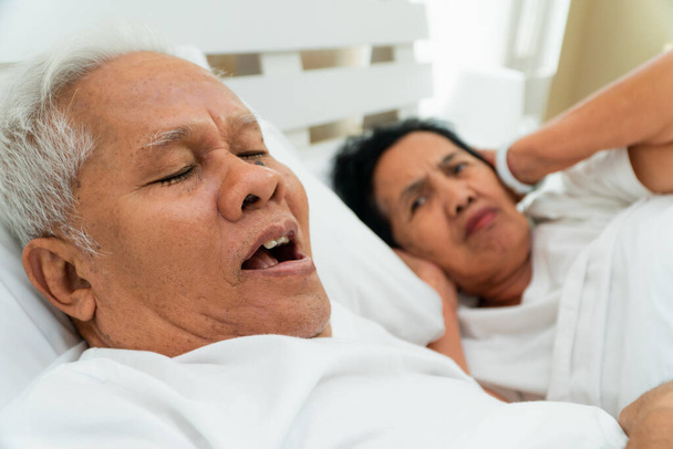 Vanhempi aasialainen nainen makaa auki silmänsä ja peittää korvansa kädet sängyssä miehensä vieressä, joka kuorsaa ja tekee melua, Siviilisäädyn ongelmia. - Valokuva, kuva