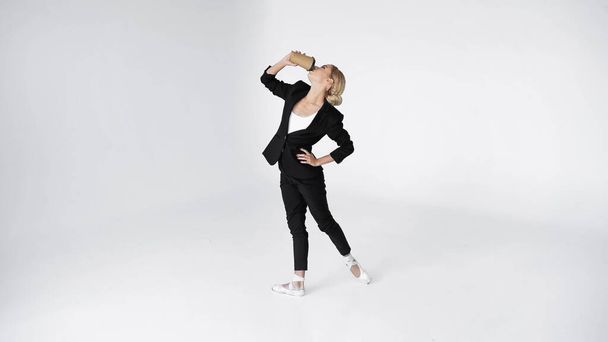 elegant ballerina in suit and pointe shoes drinking coffee to go while standing with hand on hip on white  - Foto, immagini