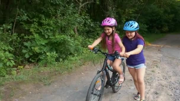 Dos chicas en cascos de bicicleta de protección en el verano en el parque. La hermana mayor ayuda a la más joven a aprender a andar en bicicleta. Feliz infancia.. - Imágenes, Vídeo