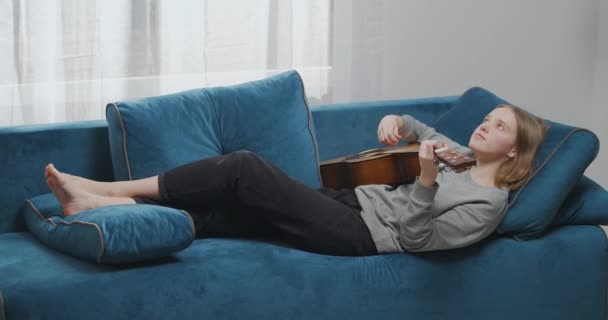 Πλευρική άποψη της χαλαρής γυναίκας που παίζει κιθάρα στο σαλόνι. Ρομαντικό κορίτσι ξαπλωμένο στον καναπέ με ένα μουσικό όργανο. Γυναικείος κιθαρίστας που ξεκουράζεται σε μαλακό καναπέ. - Πλάνα, βίντεο
